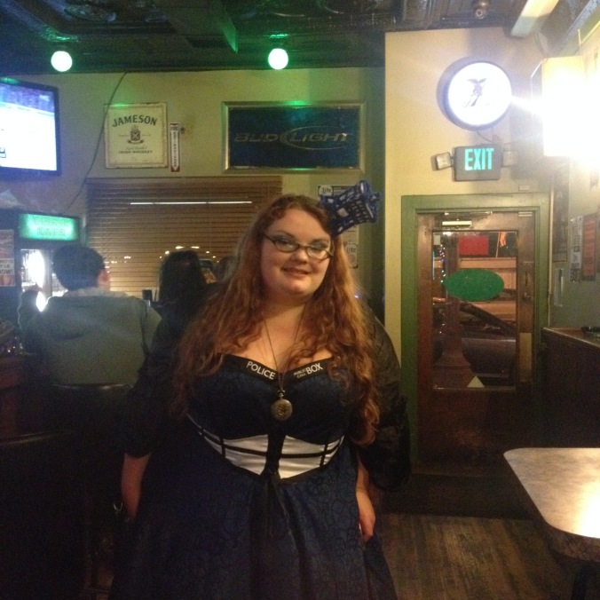 Heavy pale red headded femme in a Tardis Dress, Tardis head band, lots of makeup, and steam-punky jewelry in a pub