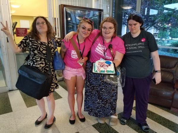 Four light skinned people. They are standing. One is holding out a cake that says, Happy Birthday ADA with the ADAPT logo, and candles spelling out 27. It's in the lobby of an apartment building somewhere in DC.