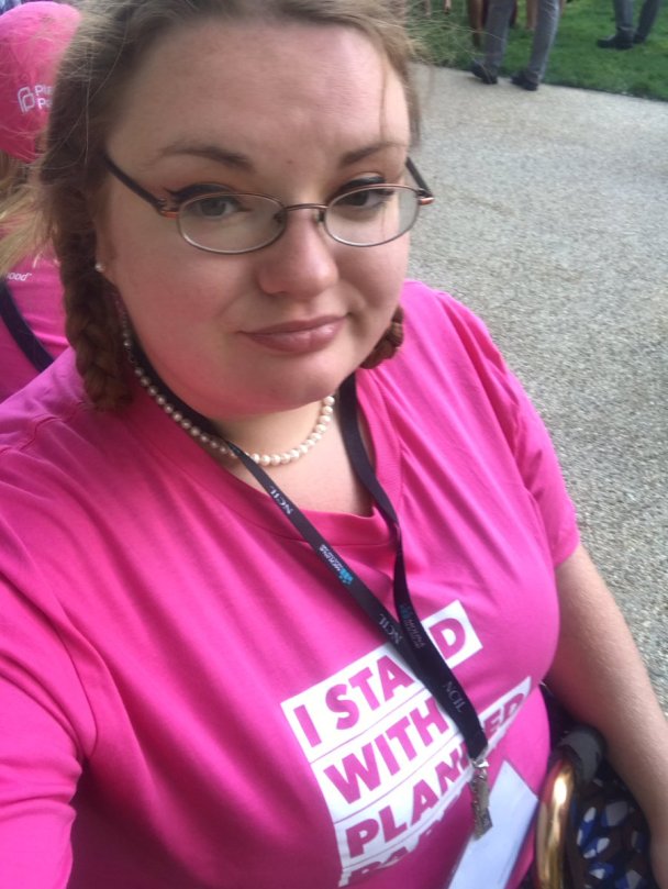 A heavy pale femme wearing pearls has on a pink "I Stand With Planned Parenthood" t-shirt on. This is a selfie.